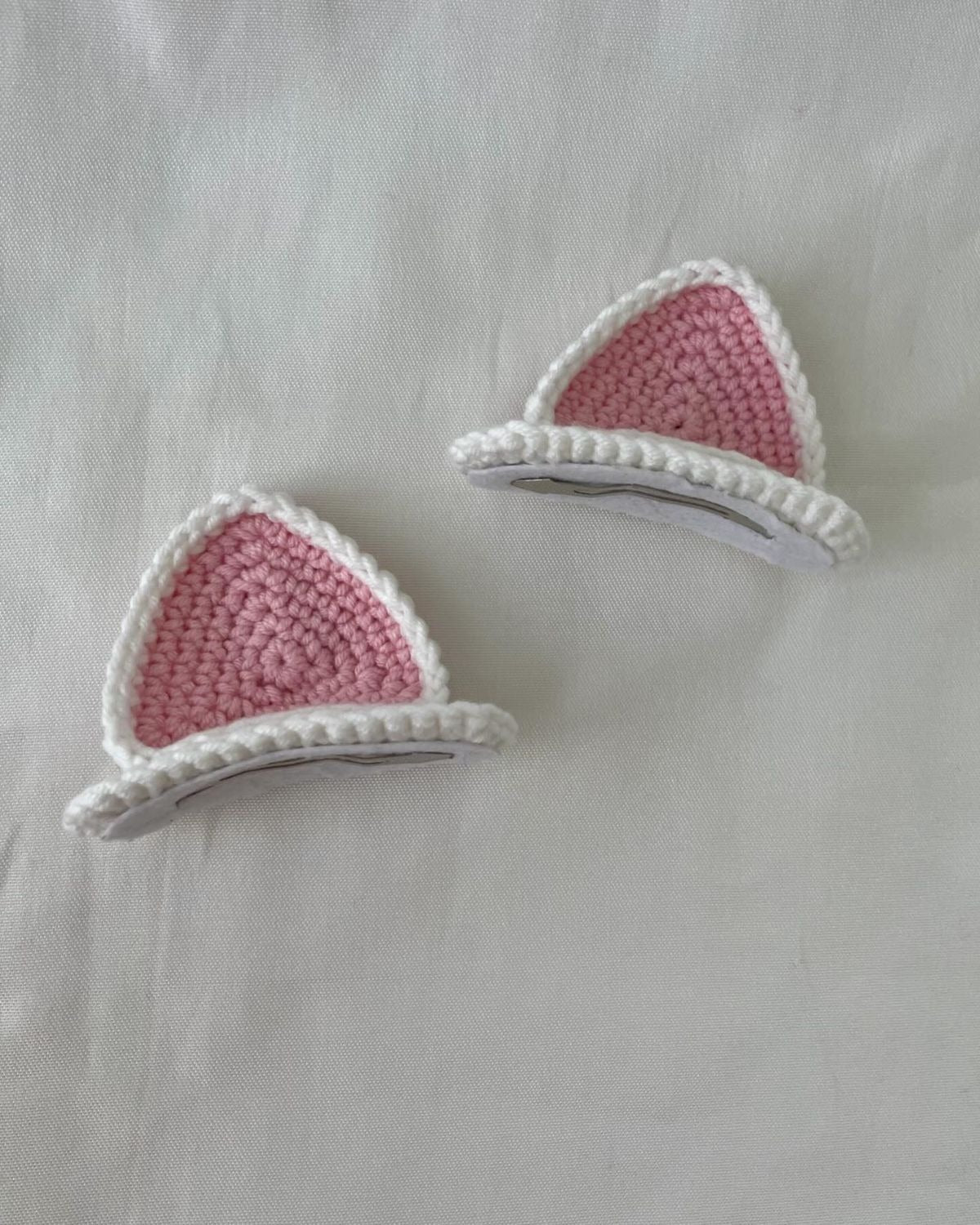 Handmade pink and white crochet cat ear hair clips set placed on white fabric background.