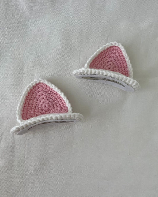 Handmade pink and white crochet cat ear hair clips set placed on white fabric background.